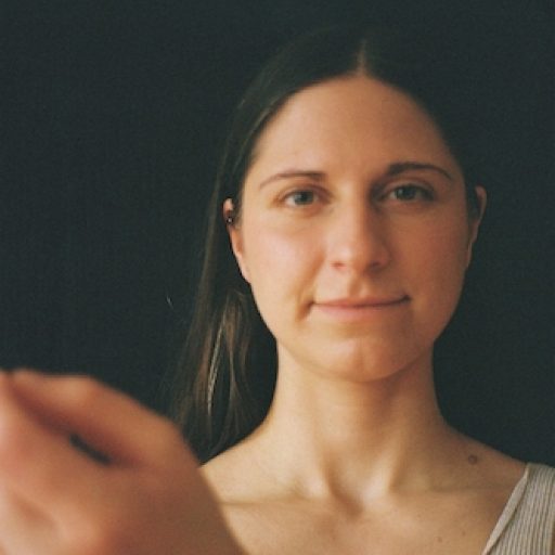 Doula und Geburtsbegleiterin in Berlin, Maria-Lisa Höcker, schaut direkt in die Kamera, eine Hand ist leicht gehoben und in der unteren linke Ecke sichtbar. Sie trägt ein beiges Oberteil vor dunklem Hintergrund