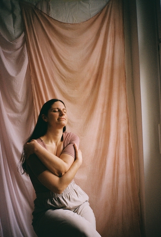 Doula in Berlin Maria-Lisa Höcker, in rosa Oberteil und beiger Hose gekleidet, sitzt vor einer Wand mit fließenden Stoffen in rosé-Tönen und umarmt sich mit beiden Armen selbst. Ihre Augen sing geschlossen.