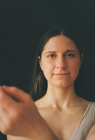 Doula und Geburtsbegleiterin in Berlin, Maria-Lisa Höcker, schaut direkt in die Kamera, eine Hand ist leicht gehoben und in der unteren linke Ecke sichtbar. Sie trägt ein beiges Oberteil vor dunklem Hintergrund