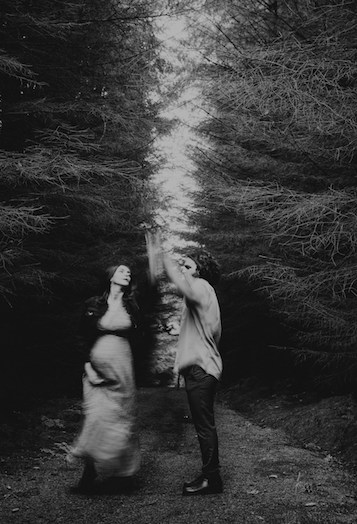 schwarz-weißes, leicht verschwommenes Bild zeigt in schwangeres Paar im Wald. Beide haben jeweils einen Arm gehoben und halten sich dort an den Händen, sie tanzen