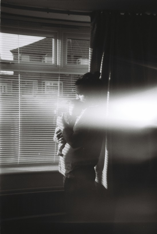 schwarz-weiß, leicht verschwommenes Bild. Ein Vater steht mit seinem Neugeborenen am Fenster und gibt ihm einen Kuss auf den Kopf.